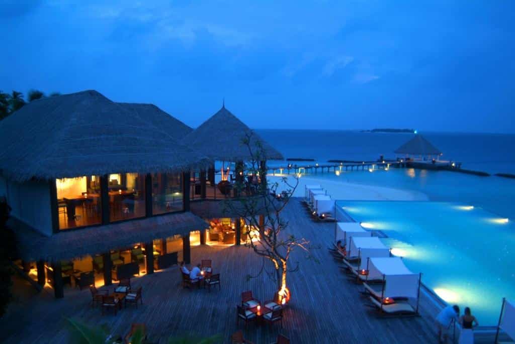 vista do Coco Bodu Hithi, um dos resorts nas Maldivas, ao entardecer, com piscina iluminada grande com vista direta para o mar e com vista também para o restaurante com várias mesinhas e cadeiras cobertas