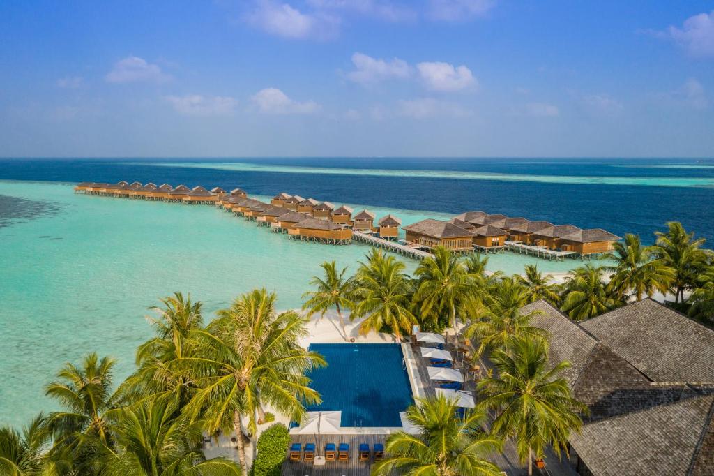vista aérea do Vilamendhoo Island Resort & Spa, um dos resorts nas Maldivas, com uma piscina retangular cercada de árvores e diversos bangalôs acima do mar mais ao longe e águas límpidas