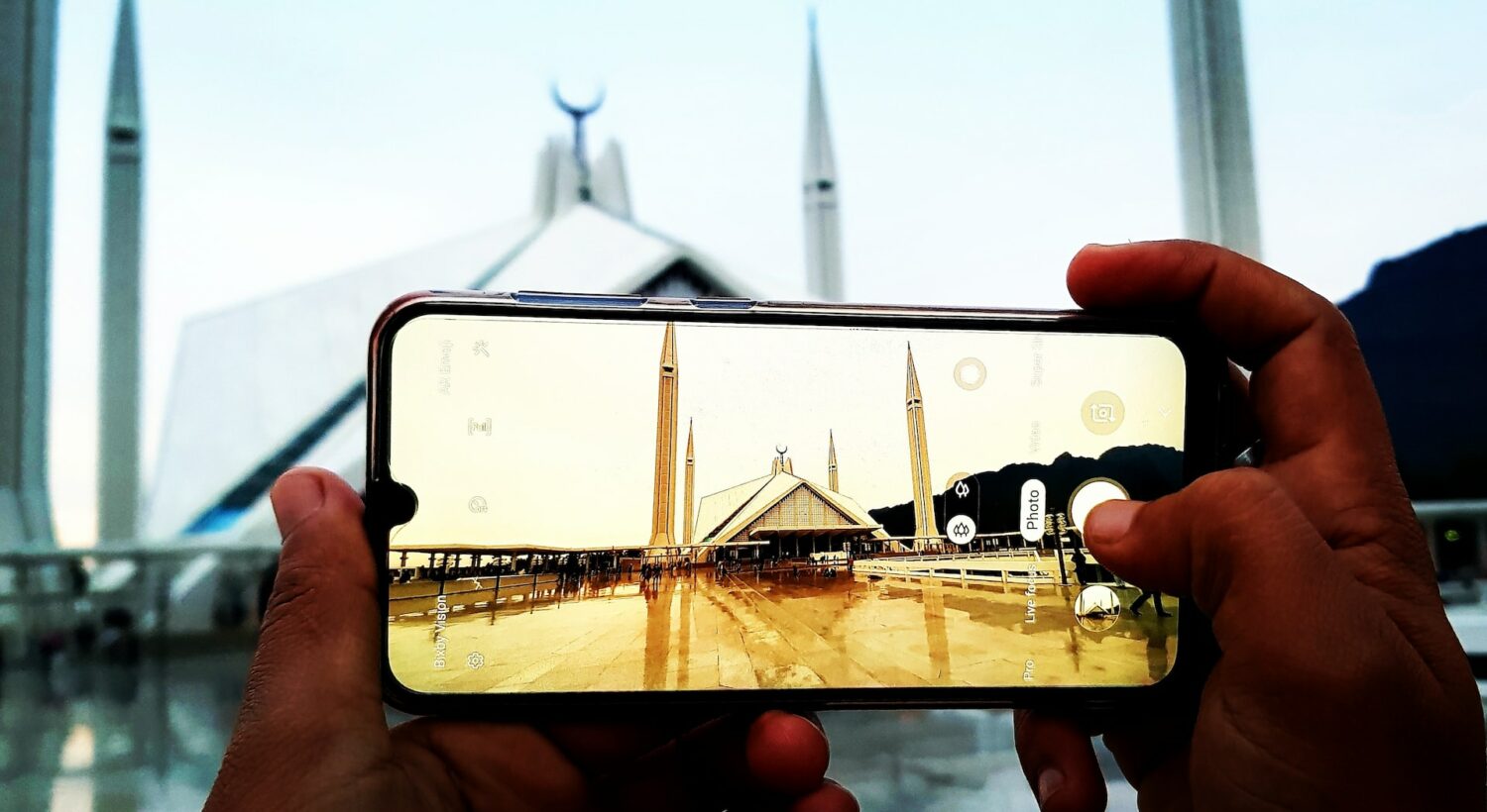 Mãos segurando um celular enquanto tira foto de uma mesquita. O tempo está nublado e o chão molhado de chuva. - Foto: Waqas Rana via Unsplash