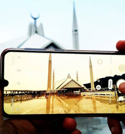 Mãos segurando um celular enquanto tira foto de uma mesquita. O tempo está nublado e o chão molhado de chuva. - Foto: Waqas Rana via Unsplash