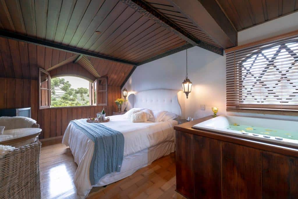 Quarto do WE Hotel com uma cama de casal, uma janela ampla, uma banheira de hidromassagem, chão de madeira e duas poltronas, para representar onde ficar em Campos do Jordão