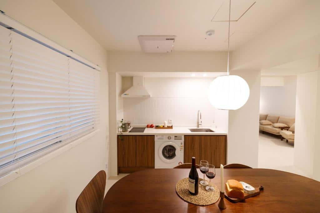 Uma pequena cozinha compacta em madeira e com itens brancos, uma mesa de refeições com seis lugares, também de madeira e uma janela com persianas brancas