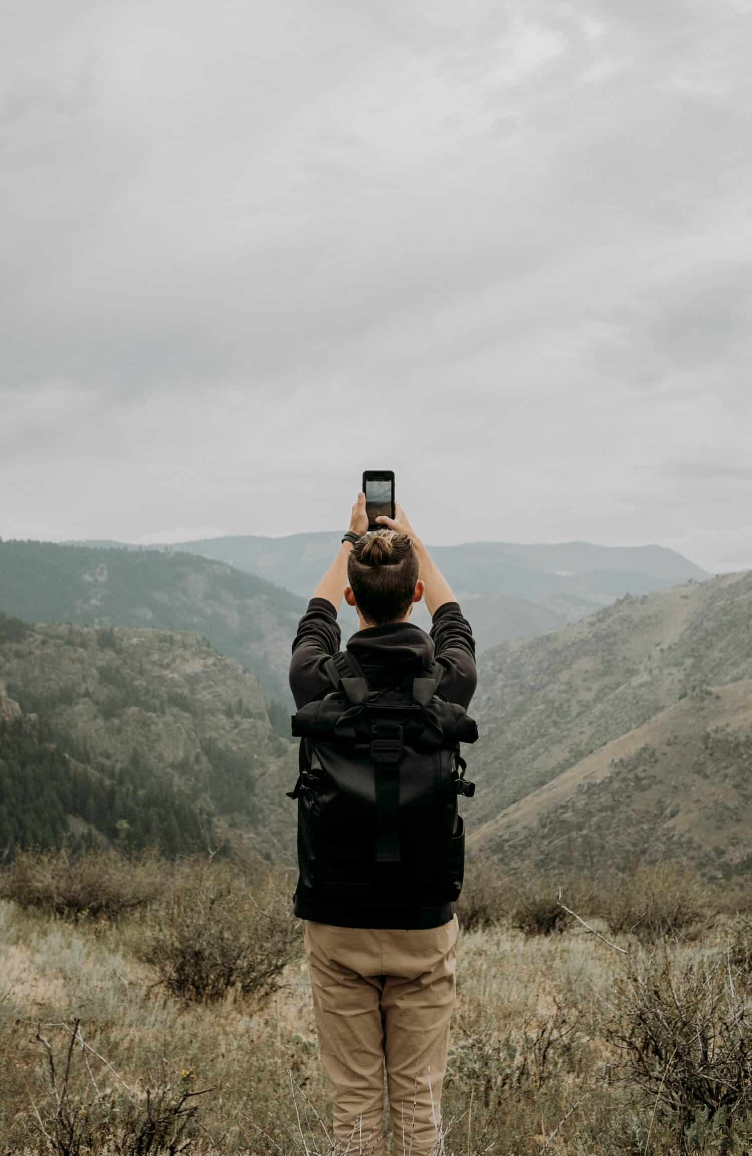 pessoa branca em meio a natureza com mochila, erguendo um celular acima da cabeça em que tira uma foto da paisagem, que é o Lookout Mountain, Denver, nos Estados Unidos, sendo um dos usos para o chip internacional pré-pago
