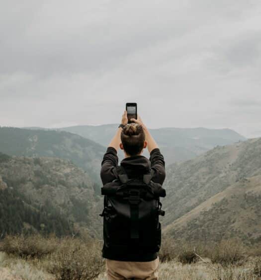 pessoa branca em meio a natureza com mochila, erguendo um celular acima da cabeça em que tira uma foto da paisagem, que é o Lookout Mountain, Denver, nos Estados Unidos, sendo um dos usos para o chip internacional pré-pago