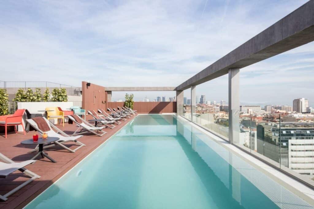 Área da piscina do Acta Voraport, uma das recomendações de hotéis baratos em Barcelona. Há espreguiçadeiras brancas ao lado da piscina, e um muto de vidro separa a piscina da beira do hotel.. É possível ver a cidade ao longo da extensão do vidro.