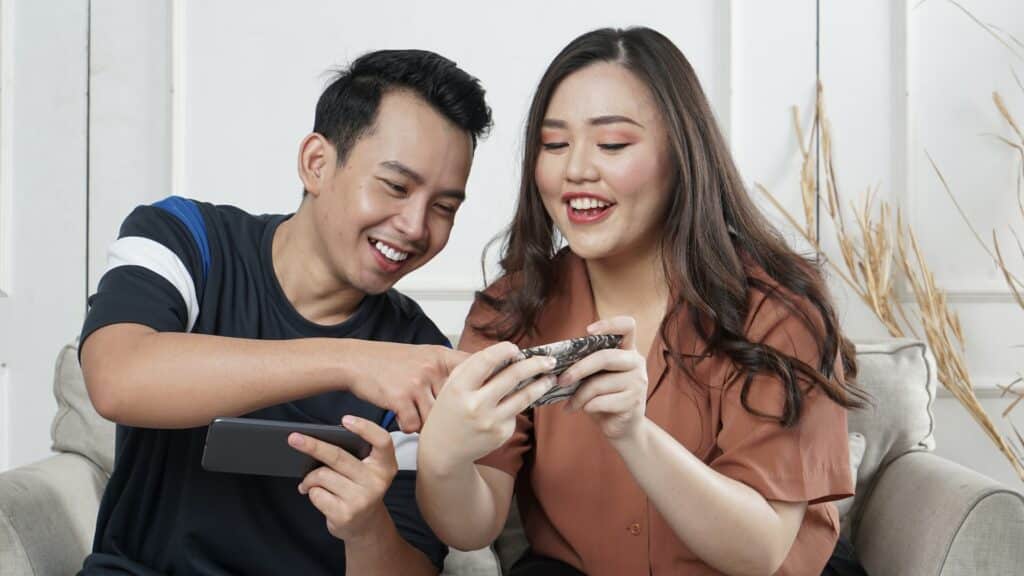 Um homem e uma mulher sentadas em um sofá, cada um com celular em mãos enquanto eles conversam e sorriem ao mexerem nos aparelhos móveis
