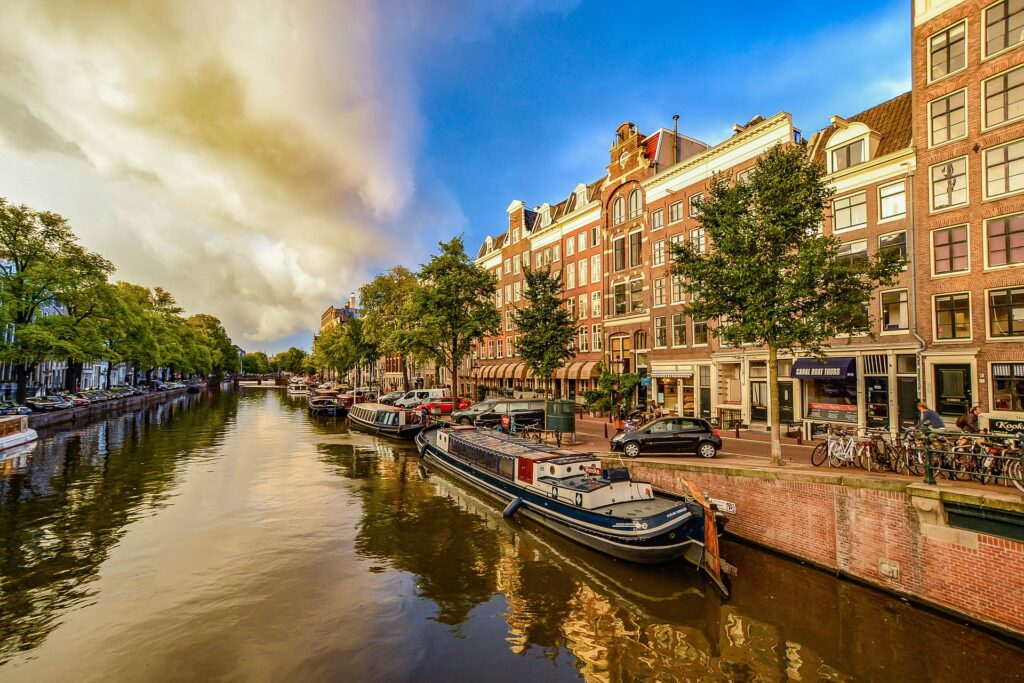 Canal de Amsterdam, com alguns barcos encostados em um muro baixo na costa, carros e bicicletas estacionadas na rua do lado, árvores e um céu azulado com nuvem branca