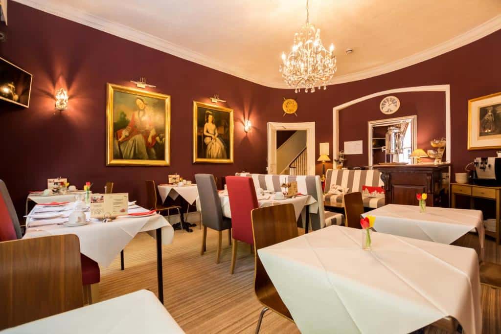 Área de refeições do Arosfa Hotel London by Compass Hospitality com chão de madeira, mesas quadradas com cadeiras estofadas ou de madeira, um lustre no centro do ambiente que é pintado em tom de vermelho escuro e com quadros nas paredes, para representar hotéis baratos em Londres
