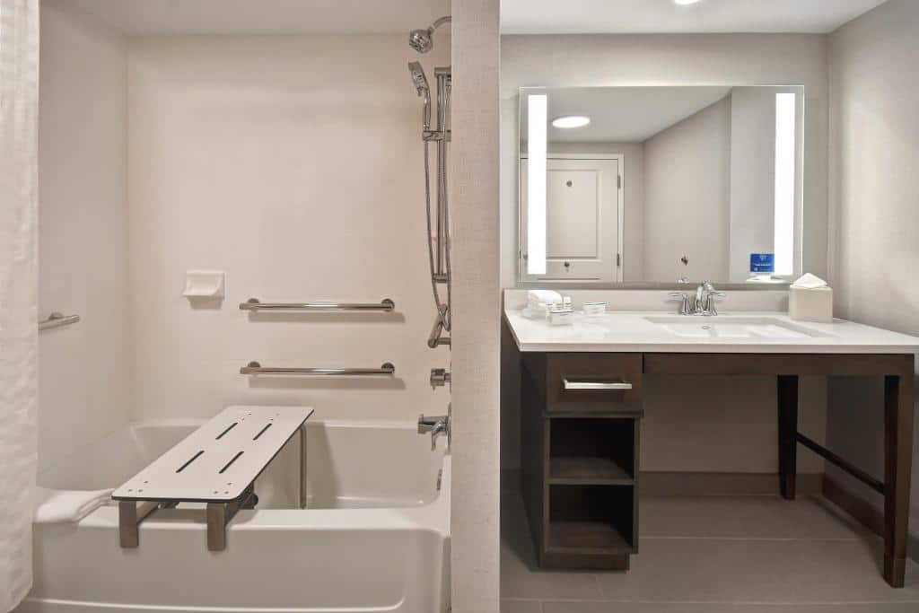 banheiro acessível do Homewood Suites By Hilton Orlando Flamingo Crossings, Fl, uma das opções de onde ficar em Orlando, com adaptações, contando com barras de apoio, cadeira de banho na banheira, mini chuveiro e pia mais baixa sem gabinete com espaço para cadeira de rodas