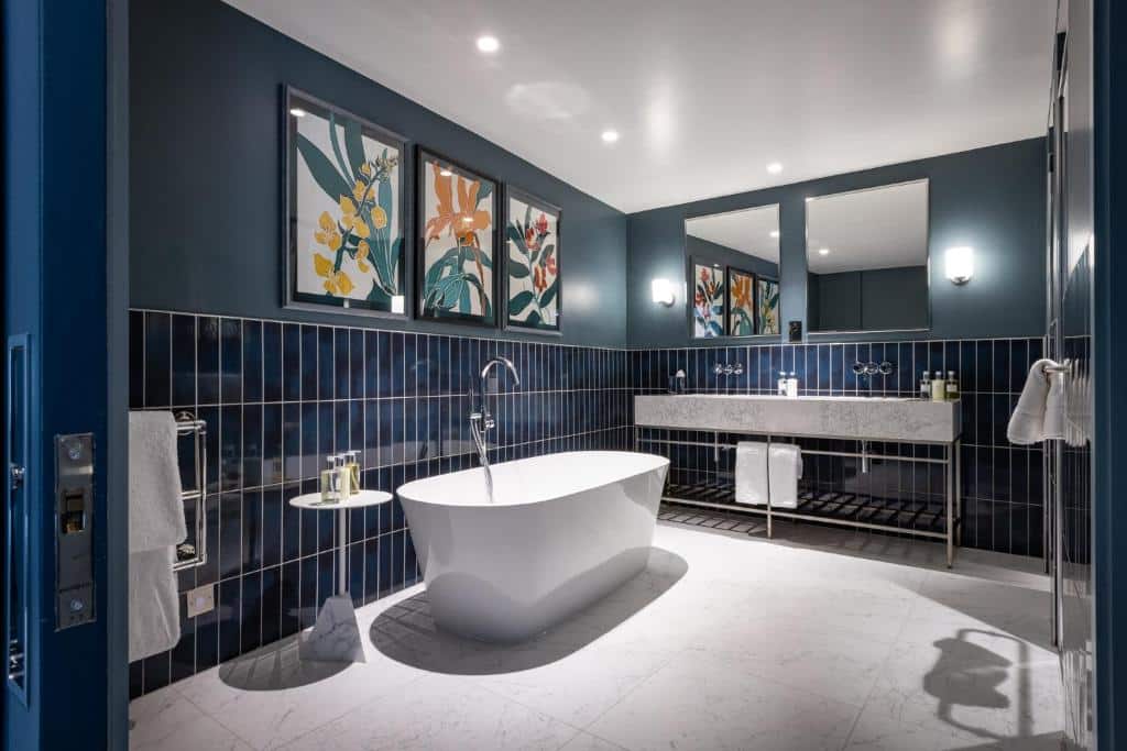 Banheiro grande do Sun Street Hotel com uma banheira oval, uma pia com duas cubas, alguns quadros na parede, dois espelhos e toalhas brancas penduradas, para representar hotéis românticos em Londres