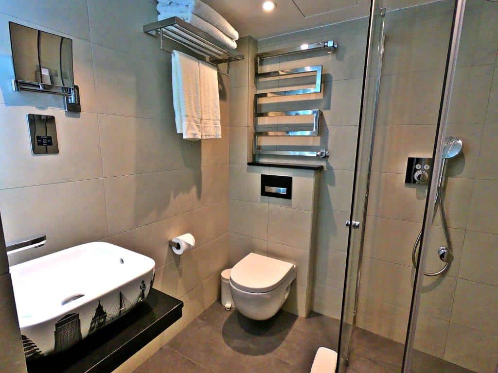 Banheiro do Best Western Plus Delmere Hotel com box de vidro, cuba branca, um pequeno espelho e barras com toalhas presas, para representar hotéis baratos em Londres