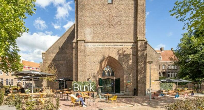 Frente do Bunk Hotel Amsterdam, um dos hotéis baratos em Amsterdam, durante o dia com o céu com poucas nuvens brancas. O prédio é histórico, feito de tijolinhos e há uma área de lazer na frente com bancos e jardim