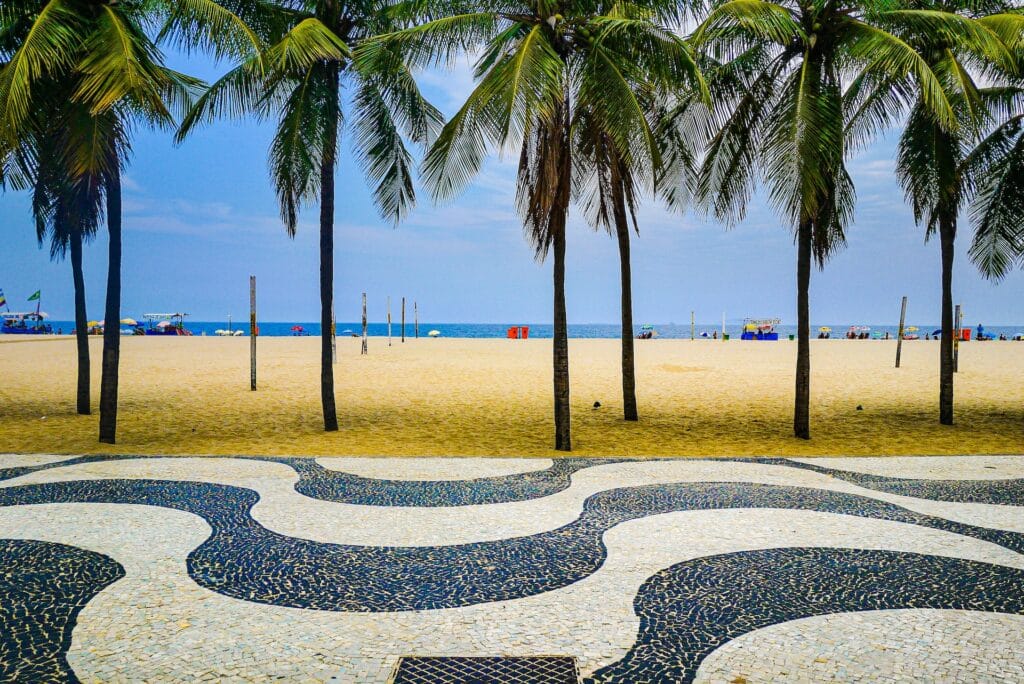 Praia de Copacabana com areia sem pessoas, alguns coqueiros, e em primeiro plano o Calçadão de Copacabana