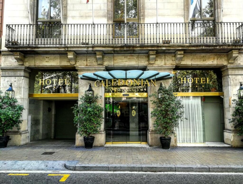 Fachada do Caledonian, uma das recomendações de hotéis em Barcelona. O nome é dourado na fachada de vidro, e há algumas plantas em frente.