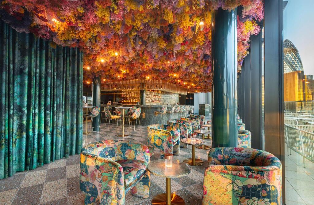 Área de refeições do Canopy by Hilton London City com janelas panorâmicas com vista para a cidade, diversas poltronas floridas, e o teto do local é cheio de flores penduradas, criando um ambiente romântico