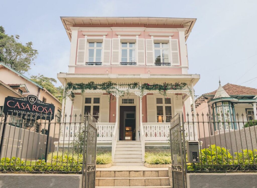 Faixada do Hotel Boutique Casa Rosa em Petrópolis, uma casa pequena com janelas e escadaria branca, paredes rosa com um letreiro e um pequeno jardim.