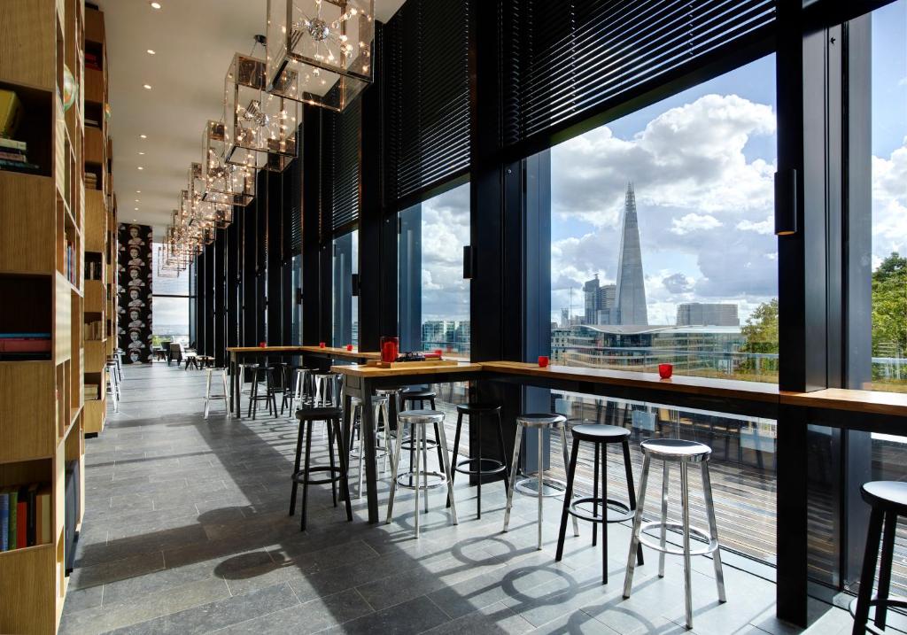 Área de refeições do citizenM Tower of London com janelas panorâmicas com vista para a cidade, diversos lustres, uma bancada de madeira com banquetas pretas para se sentar, para representar os melhores hotéis em Londres