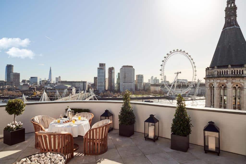 Terraço do Corinthia London com vista para a London Eye, o rio Tâmisa e a cidade de Londres, no local há uma mesa redonda com cadeiras de madeira, além de alguns vasos de plantas, para representar hotéis perto da London Eye em Londres