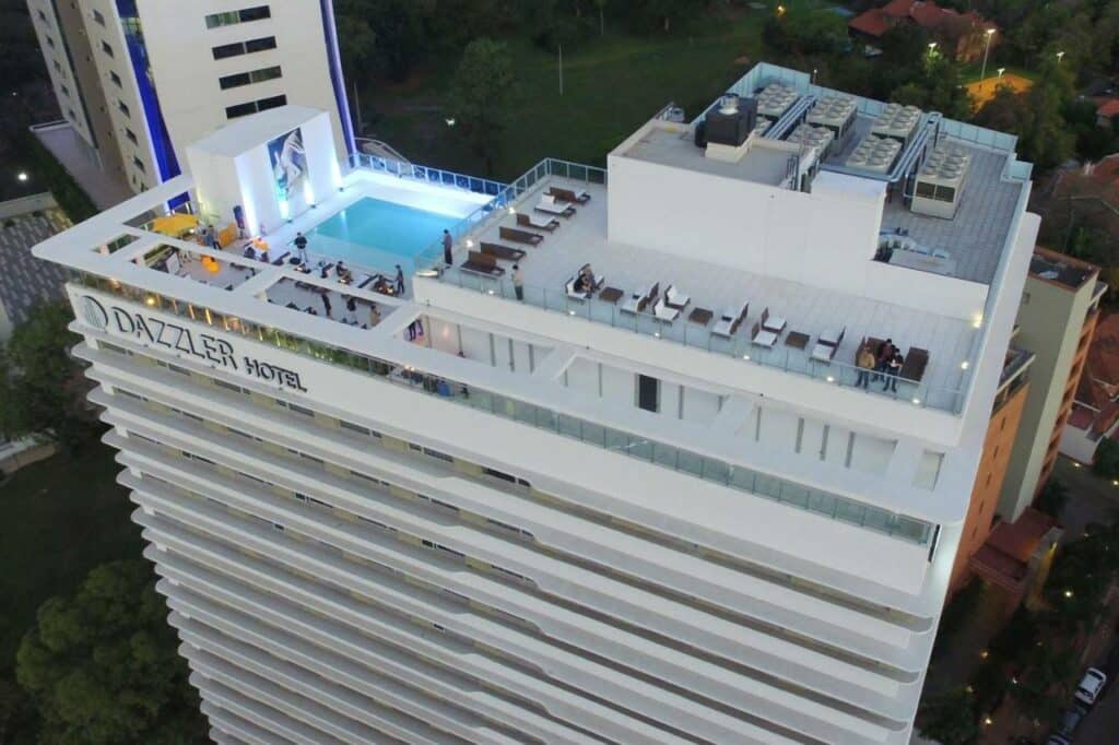 Faixada do Hotel Dazzler em Assunção no Paraguai. Hotel grande com piscina no topo, com cadeiras e algumas pessoas.