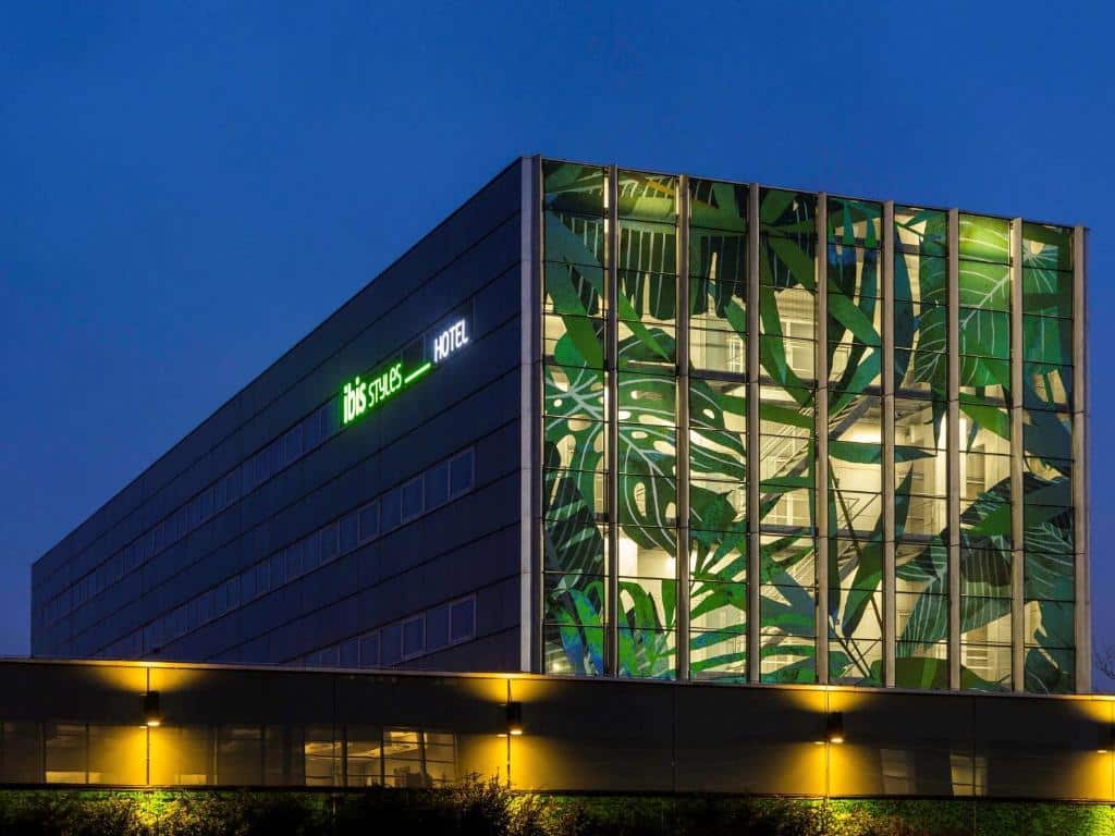 Fachada iluminada do ibis Styles Amsterdam Airport, um dos hotéis baratos em Amsterdam, durante uma noite azulada, com luzes e prédio com a frente com desenhos de folhas verdes e letreiro iluminado escrito "Ibis styles_hotel"