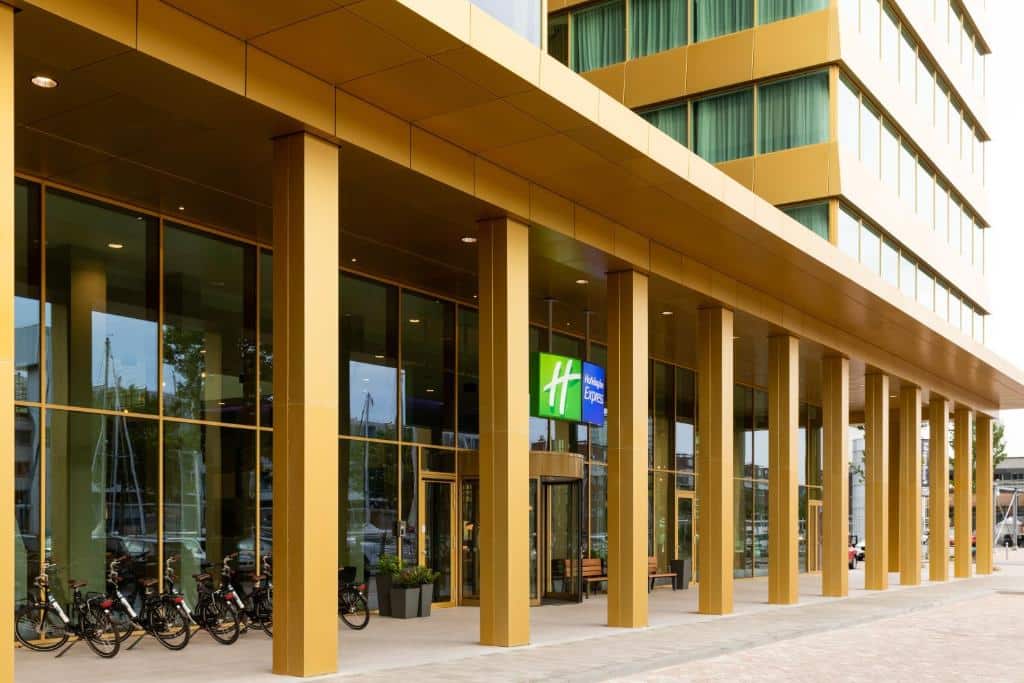 Entrada do Holiday Inn Express Amsterdam, com paredes de vidro e colunas douradas. A cima da porta de entrada há uma placa iluminada na cor verde e azul escrito "H" e "Holiday Inn Express"