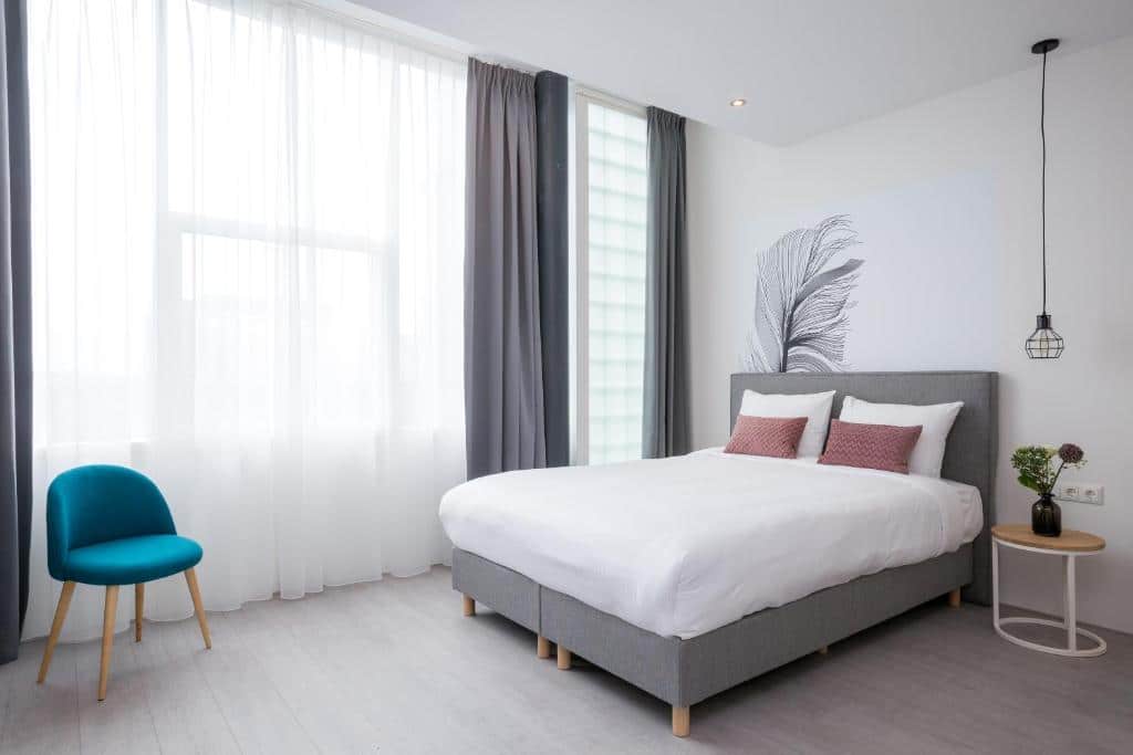 Quarto duplo comfort do Hotel2Stay, de 30 m², com cama de casal, uma poltrona azul, mesinha redonda ao lado da cama com um vaso de flor em cima e janela com cortinas brancas e cinza