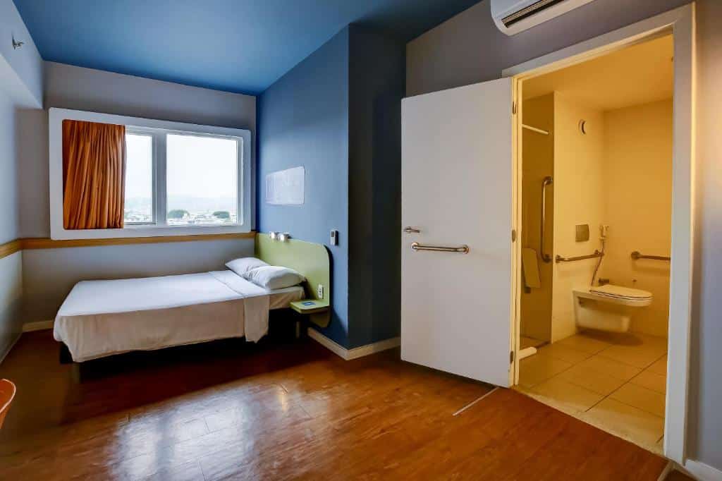 Quarto do hotel mostrando a acessibilidade na cama de casal mais baixa, porta para o banheiro mais ampla e o banheiro com recursos de acessibilidade.