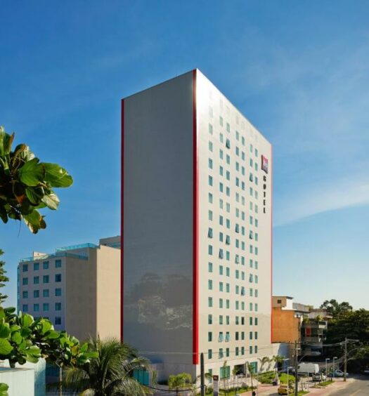 Grande prédio cinza com detalhes em vermelho, pequenos prédios ao lado e uma árvore verde durante o dia, ilustrando post hotéis Ibis no Rio de Janeiro.