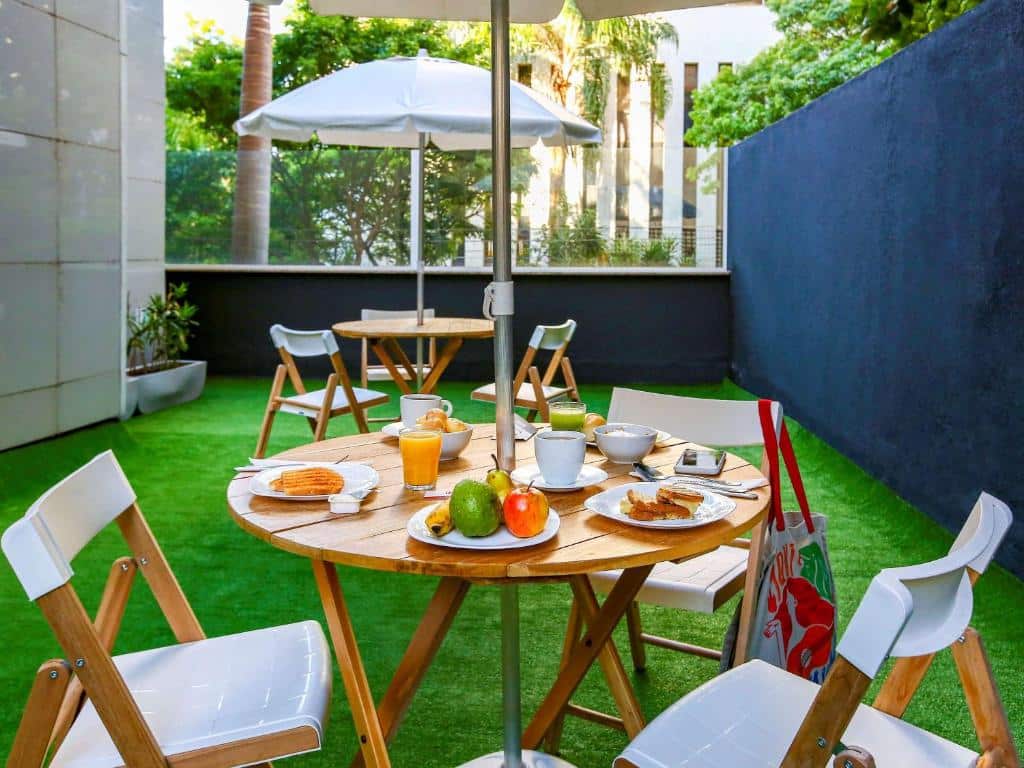 Mesa redonda de madeira e algumas cadeiras brancas em cima de um gramado verde com o café da manhã.