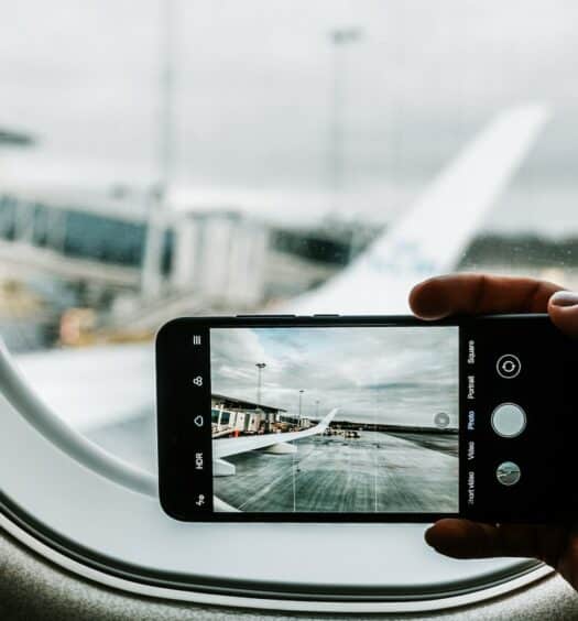 Uma mão segurando um celular na horizontal dentro de um avião, a câmera do aparelho está fotografando uma das asas do avião, para representar chip celular no Aeroporto de Guarulhos
