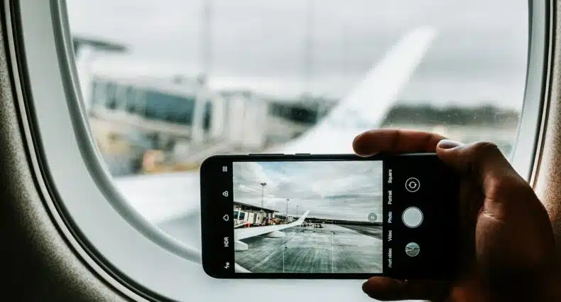 Uma mão segurando um celular na horizontal dentro de um avião, a câmera do aparelho está fotografando uma das asas do avião, para representar chip celular no Aeroporto de Guarulhos