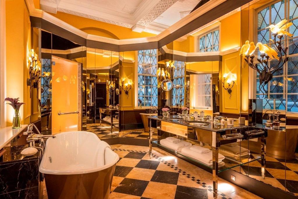 Banheiro do L'oscar London com uma banheira, uma pia com toalhas presas, alguns lustres e janelas de vidro, com piso de mármore e uma porta de vidro que leva para o box