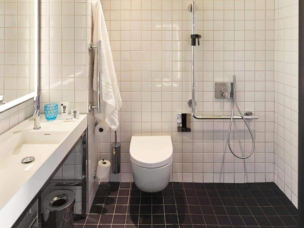 Banheiro adaptado do Mama Shelter London – Shoreditch com um vaso sanitário com barras de apoio, um box sem vidro com barras de apoio e uma pia mais baixa, para representar hotéis baratos em Londres