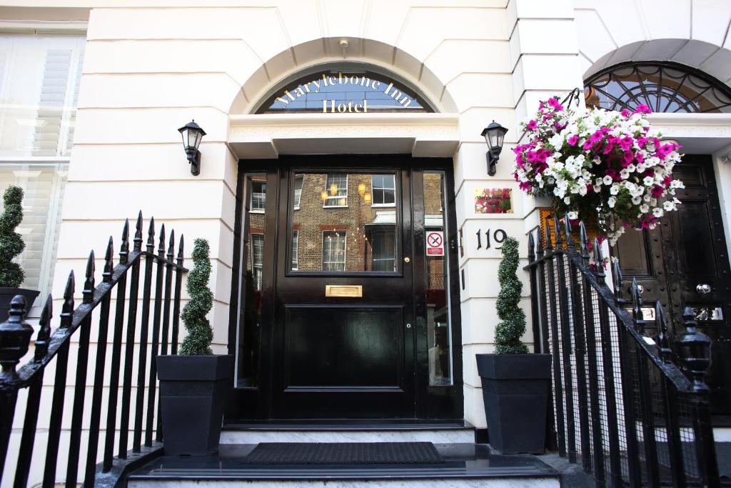 Entrada do Marylebone Inn com uma ampla porta preta com vidro, durante a escada há enfeitas com flores nos dois corrimãos, a construção é em estilo europeu e em tons de branco