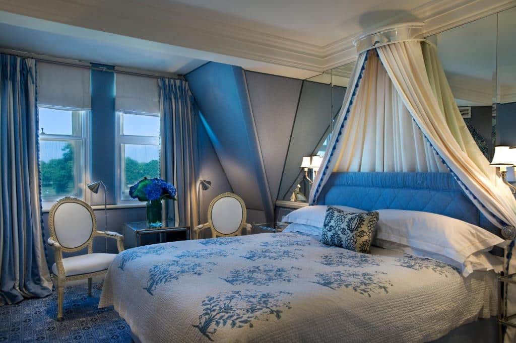 Quarto do Milestone Hotel Kensington em estilo vitoriano, tudo em branco e azul, com uma cama de casal, uma janela com cortinas, duas cadeiras, e um espelho na cabeceira da cama, para representar os melhores hotéis em Londres