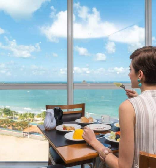 Mulher de cabelo curto, sentada em frente a uma mesa com café da manhã, com uma das mãos segurando um garfo com fruta perto da boca, olhando para paredes de vidro do ambiente com vista panorâmica do mar. Representa hotéis em Maceió