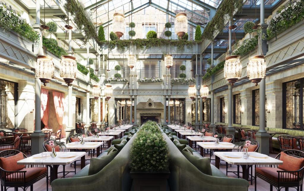 Área aberta do NoMad London  com um teto de vidro muito alto cercado por plantas e luminárias penduradas, há sofás verdes e mesas com cadeiras de madeira, para representar hotéis românticos em Londres