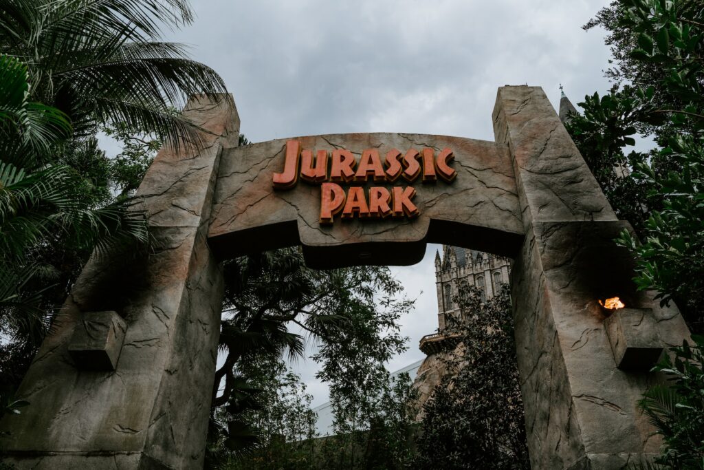 entrada com duas torres de "pedra" ligadas com os escritos em vermelho do Parque temático Jurrasic Park na Universal, em Orlando, com diversas árvores ao redor
