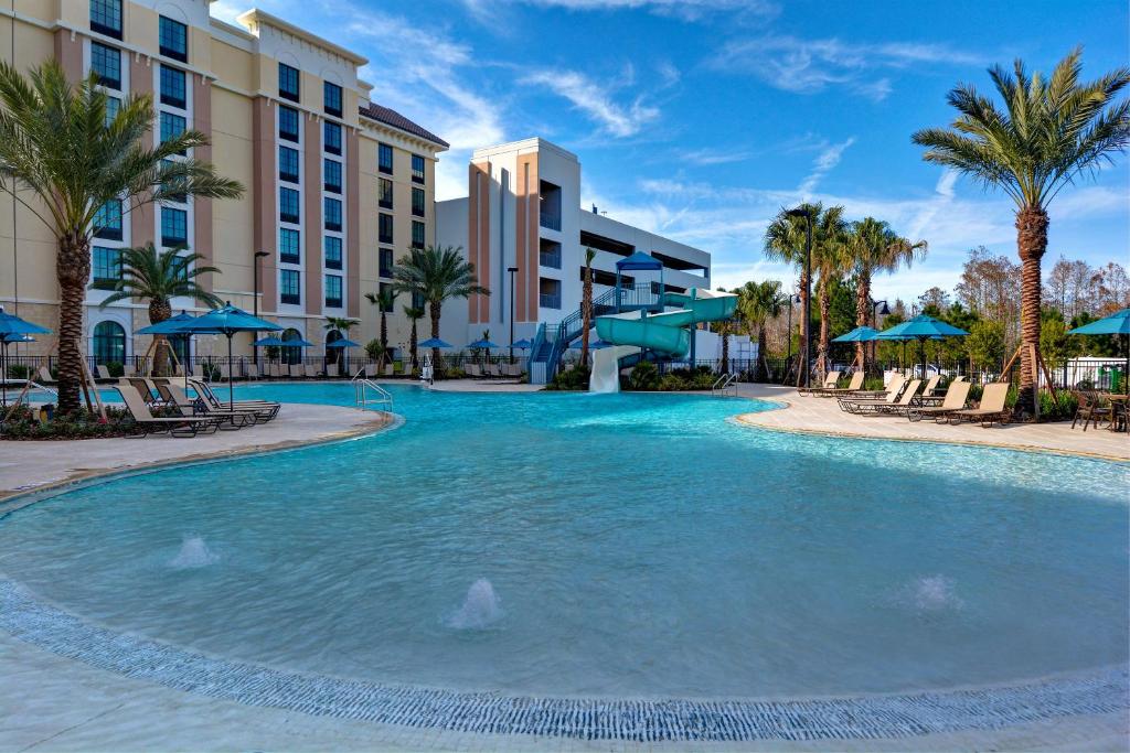 piscina ampla com bordas sinuosas com espreguiçadeiras ao redor com palmeiras e a estrutura do Home2 Suites By Hilton Orlando Flamingo Crossings, FL atrás, sendo uma das opções de onde ficar em Orlando
