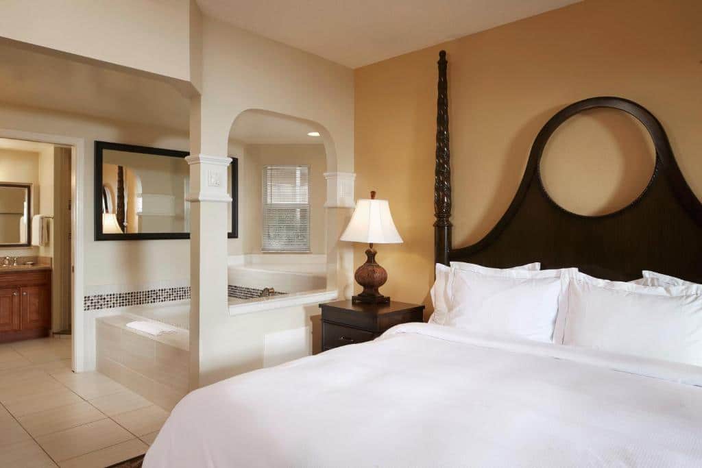 quarto com hidro do Hilton Grand Vacations Club SeaWorld Orlando com cama de casal grande, cabeceira arredondada com mesinha e luminária ao lado e com uma hidro ao lado em uma parede com uma parte aberta