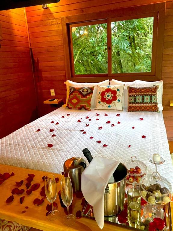 quarto do Bangalô dos Alpes de São Chico com vista para a cama enfeitada com três almofadas estampadas, pétalas de rosas vermelhas jogadas sobre a colcha e um balde de inox na bancada em frente a cama com champanhe, taças de vidro, morangos e chocolates