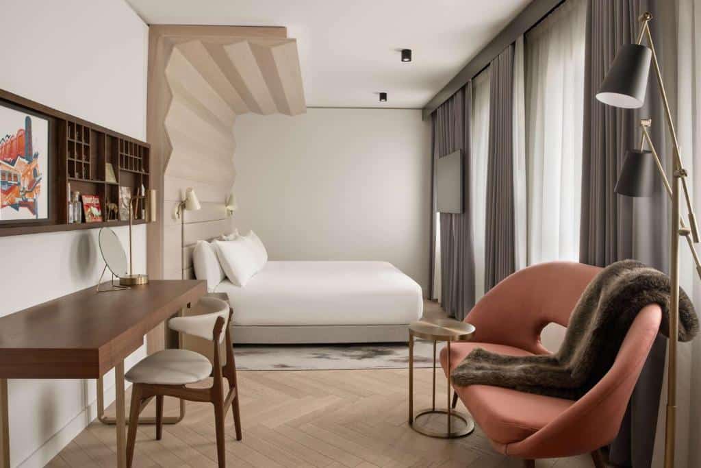 Quarto do Canopy by Hilton London City com uma cama de casal, uma cabeceira que vai até o teto, uma poltrona grande, duas janelas com cortinas, uma televisão, uma mesa com uma cadeira e o chão é de madeira