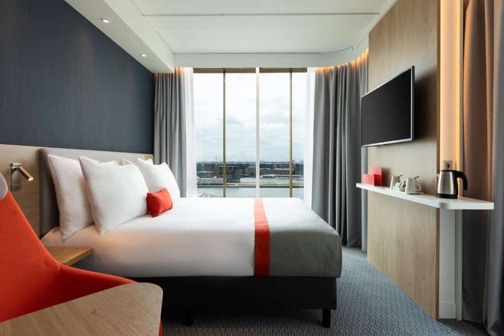 Quarto duplo standard do Holiday Inn Express Amsterdam, de 23 m², com chão de carpete, uma cama de casal com travesseiros em cima, TV, uma prateleira com utensílios para fazer chá ou café e uma janela panorâmica de vidro com vista do canal e dos prédios