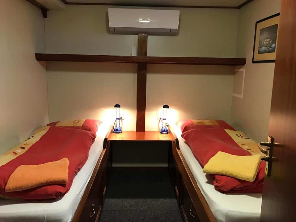 Quarto do Hotelboat Angeline, de 10 m², com duas camas de solteiro com cobertores e travesseiros em cima e uma mesinha de madeira ao meio com abajures. Há também ar-condicionado e uma prateleira de madeira