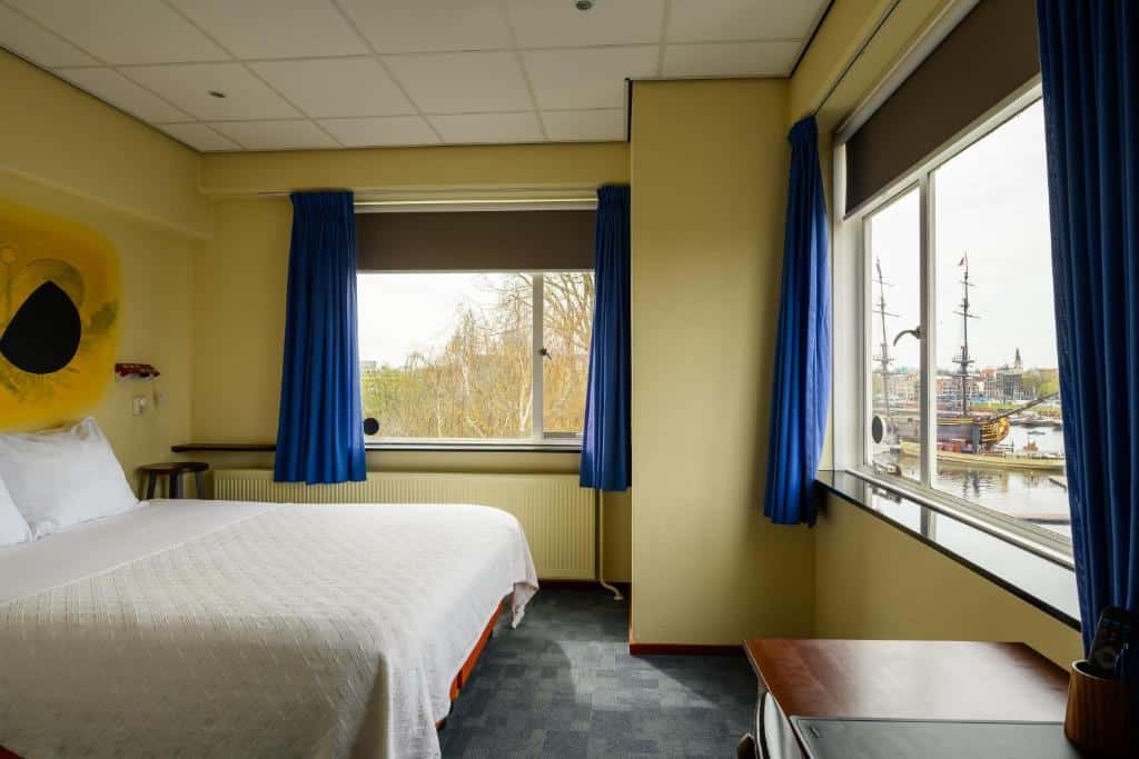 Quarto com cama de casal do Pension Homeland, de 17 m², com chão de carpete, paredes amarelo claro e duas janelas abertas com cortina azul aberta e vista de árvores e canal com barco no lado de fora