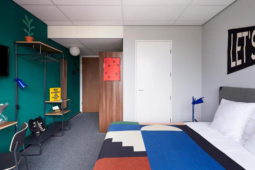 Estúdio duplo deluxe do The Social Hub Amsterdam West 4 star, de 21 m², com chão de carpete cinza escuro, parede verde escuro no lado esquerdo com cabideiro e mesinhas na frente, e no lado direito tem uma cama com lençol colorido e uma porta branca fechada ao fundo