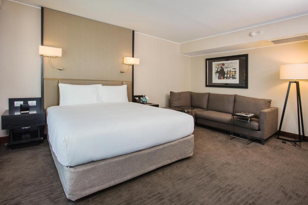 Quarto do DoubleTree by Hilton com cama de casal no meio do quarto com duas cômodas ao lado e, do lado esquerdo, um sofá de canto.