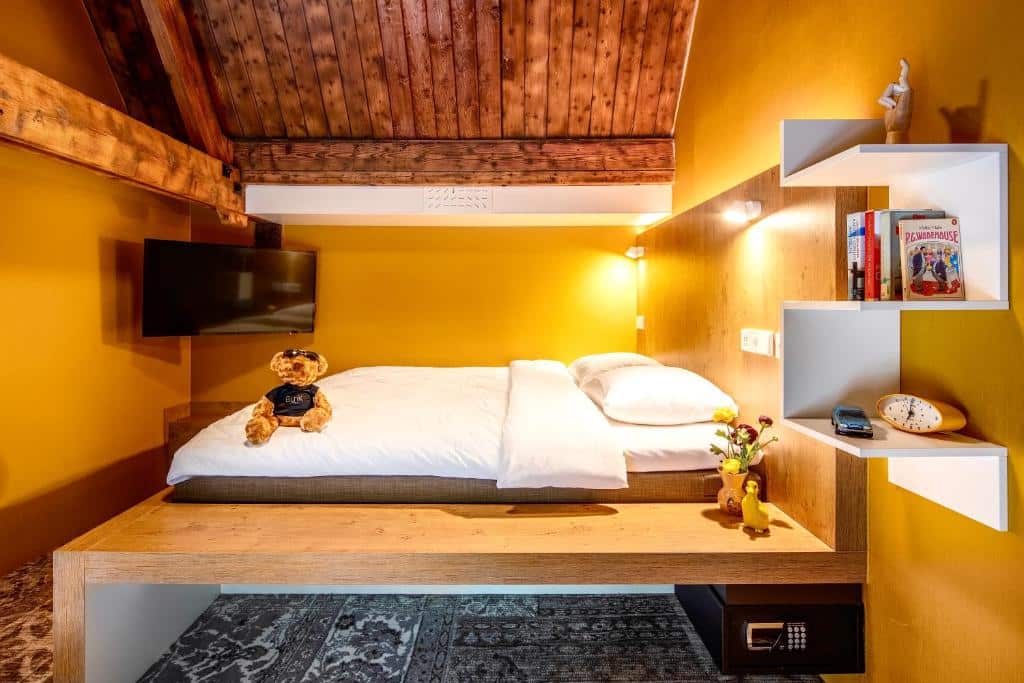 Quarto epic do Bunk Hotel, de 16 m², com uma cama com base de madeira, TV, cofre abaixo da cama, teto de madeira e paredes amarelo mostarda