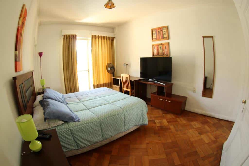 Quarto do Hostal del Cerro com cama de casal no centro, do lado da cama duas cômodas de madeira com luminária e, em frente a cama, tem uma cômoda de madeira com TV e uma mesa de trabalho.