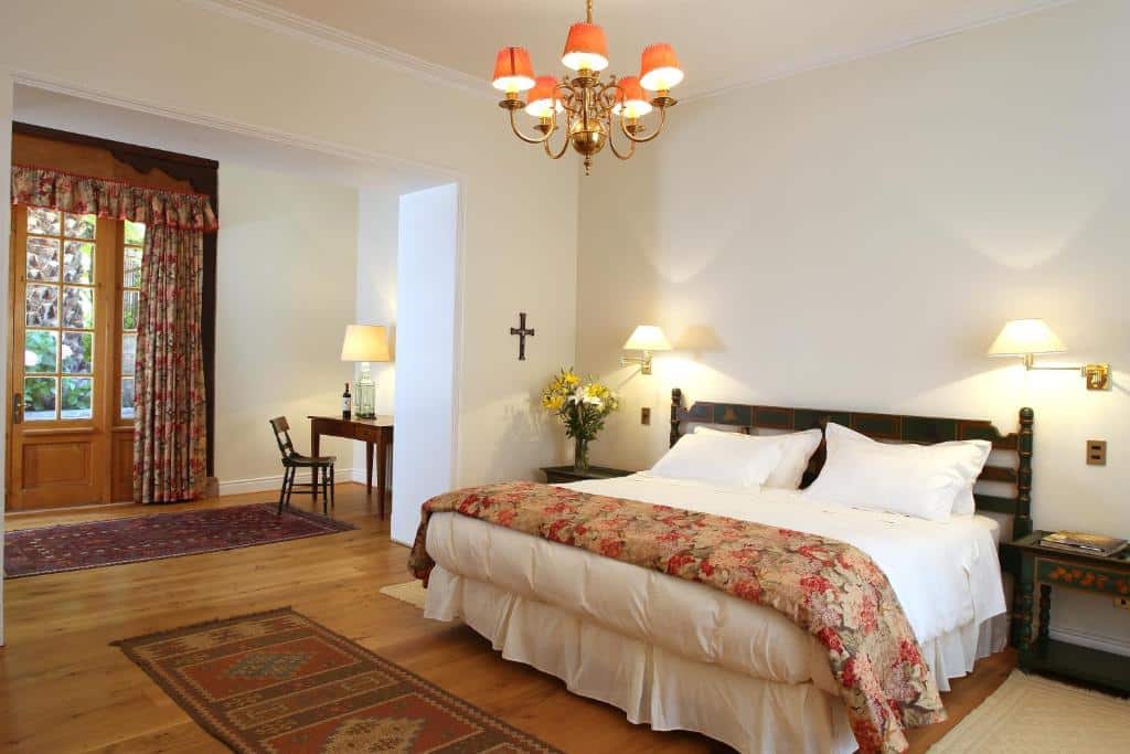 Quarto do Hotel Casa Real – Viña Santa Rita com cama de casal no meio do quarto, duas cômodas de madeira de cada lado da cama, ao fundo, uma mesa de trabalho de madeira com cadeira.